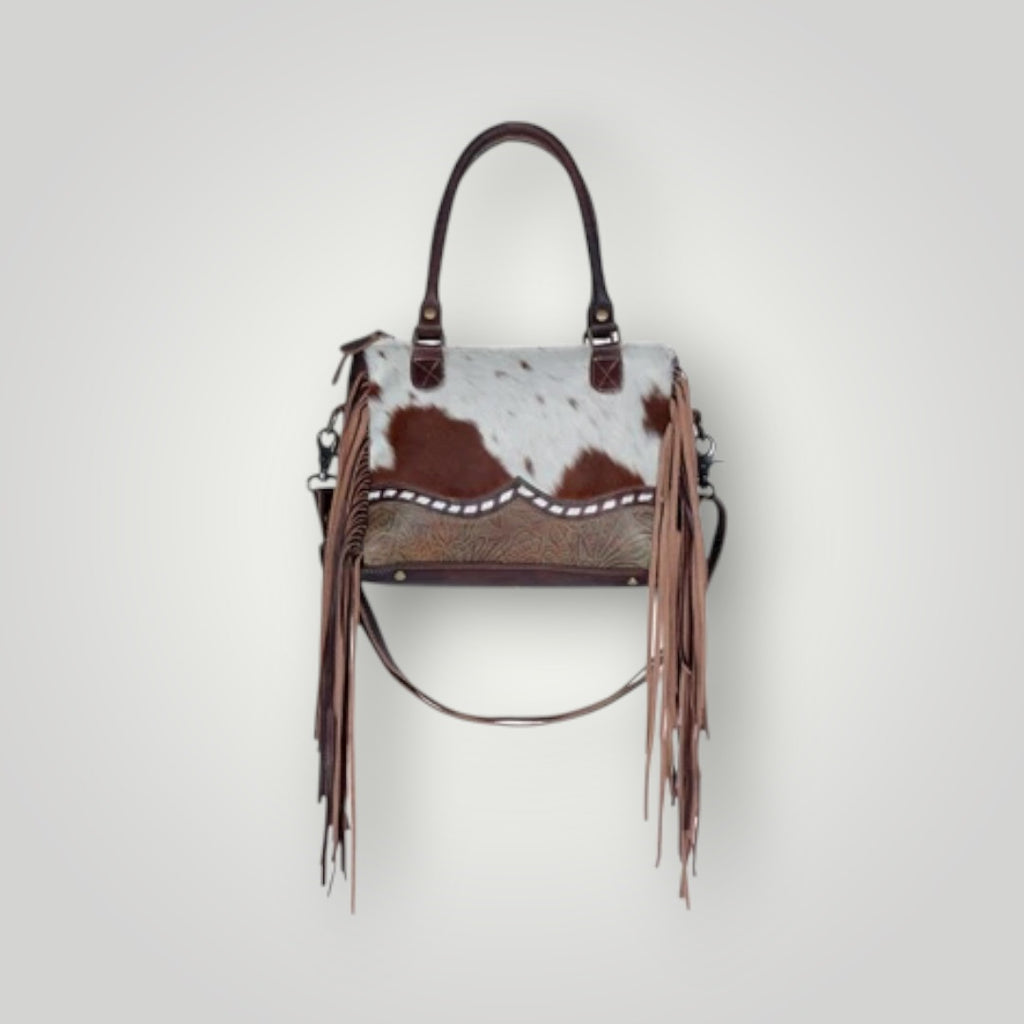 A western-style handbag featuring a brown and white cowhide patterned body, long brown leather fringe hanging from the sides, and dark brown leather top handles, shown against a light gray background.