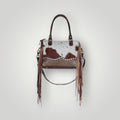 A western-style handbag featuring a brown and white cowhide patterned body, long brown leather fringe hanging from the sides, and dark brown leather top handles, shown against a light gray background.