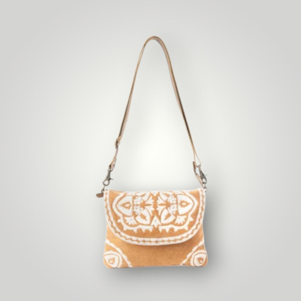 A small shoulder bag with a tan and white ikat pattern, featuring a long tan strap and silver-tone hardware, shown against a light gray gradient background.