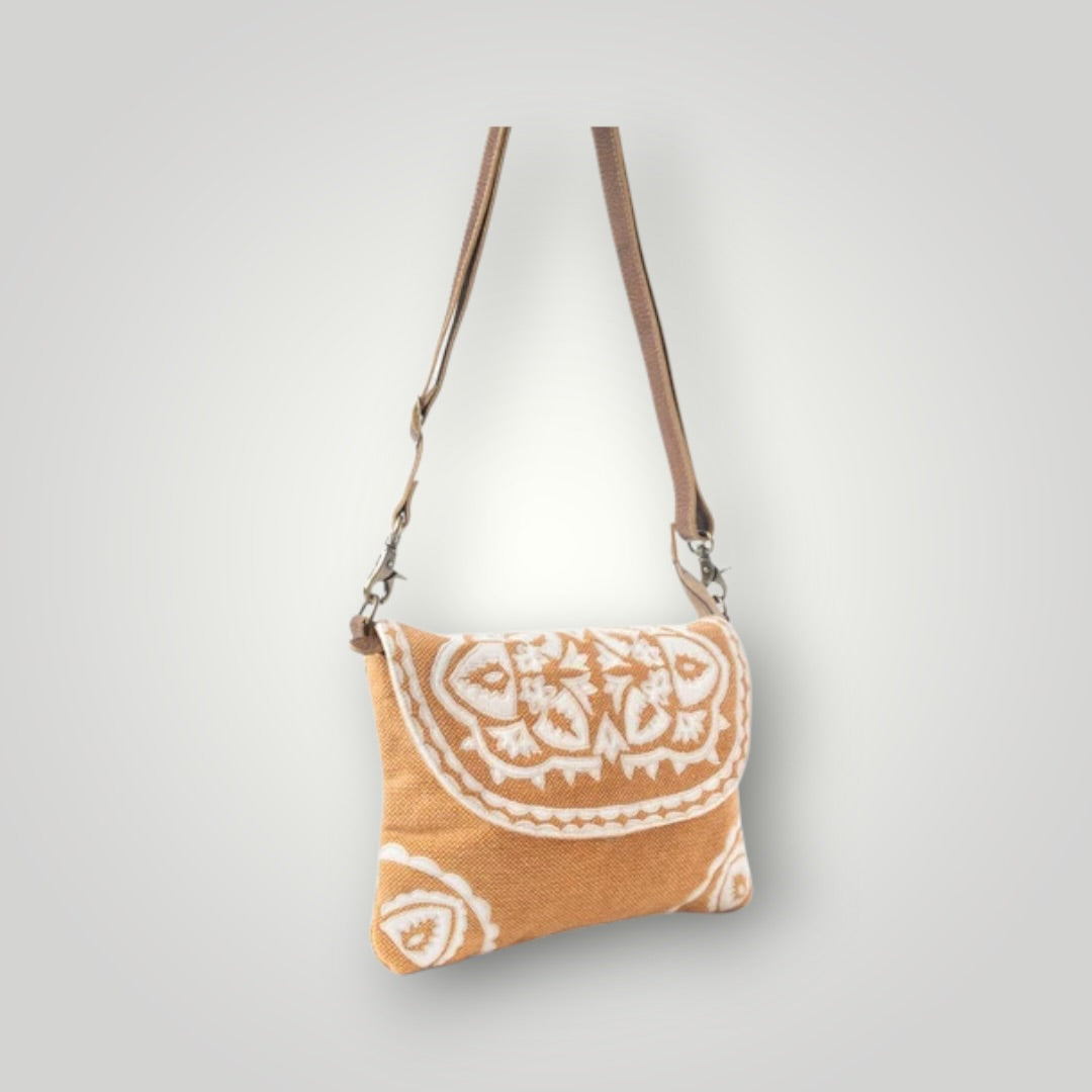 A small shoulder bag with a tan woven fabric body featuring a white hand-drawn mandala pattern on the front flap, complete with a brown leather strap and silver-tone hardware, shown against a light gray gradient background.