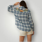 A woman shown from the back wearing a blue and white plaid button-up shirt with colorful floral embroidery across the shoulders, paired with off-white shorts, against a light gray studio background.