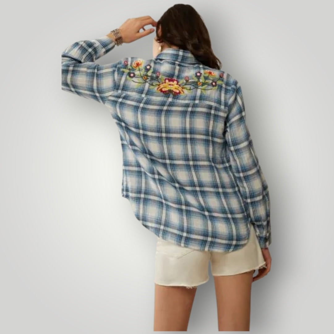 A woman shown from the back wearing a blue and white plaid button-up shirt with colorful floral embroidery across the shoulders, paired with off-white shorts, against a light gray studio background.