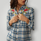 A woman wearing a blue and white plaid button-up shirt with floral embroidery on the chest, shown from the shoulders to the waist against a light gray background.