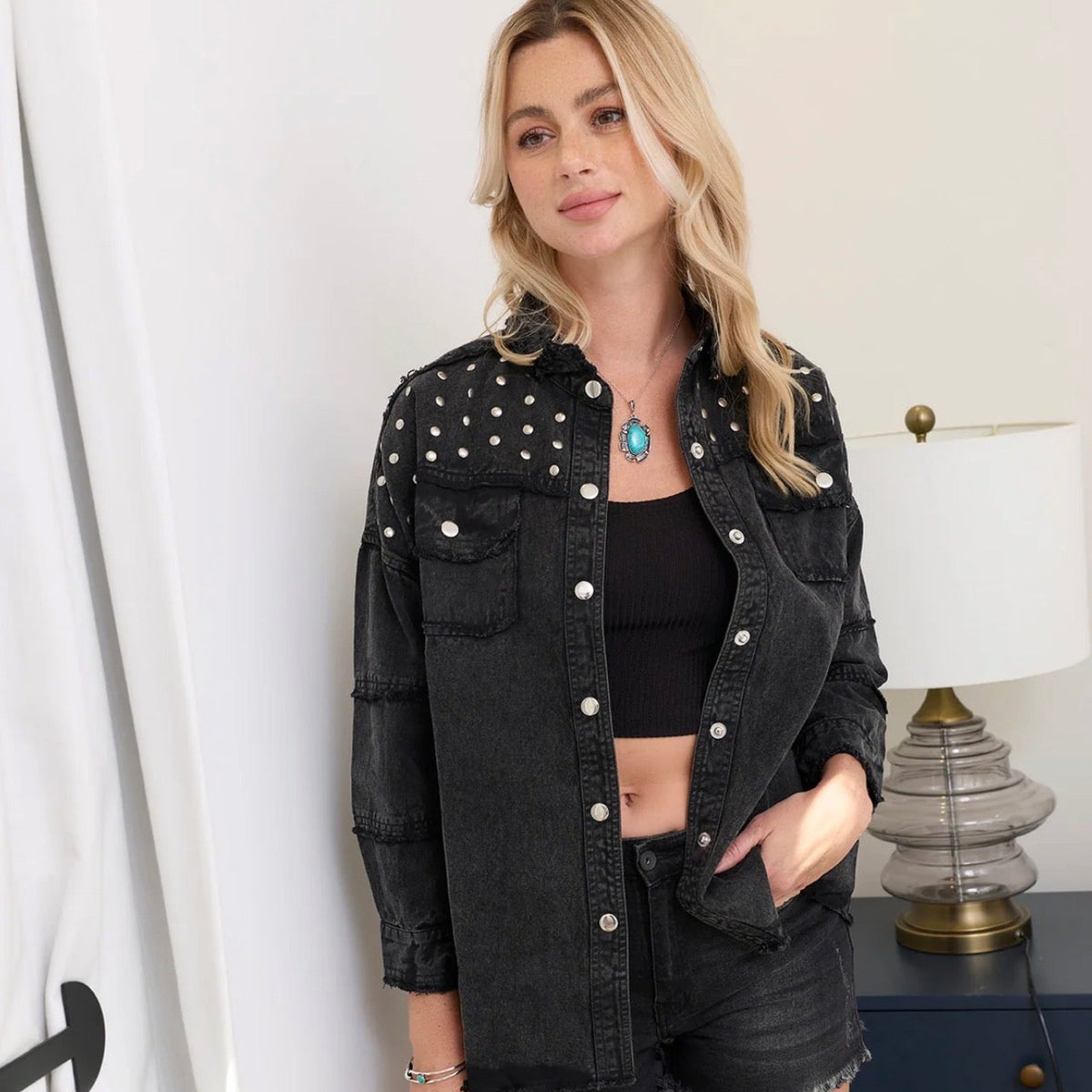 A woman modeling a black denim jacket with silver studs and silver buttons, worn over a black crop top and black denim shorts in an indoor setting.