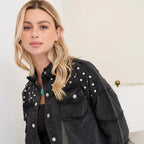 A blonde woman wearing a black washed denim jacket with silver studs and buttons, styled over a black top, in a brightly lit indoor setting.