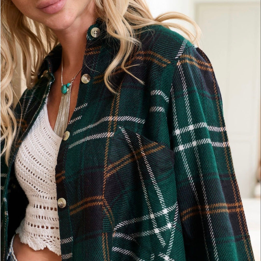 A close-up of a woman wearing a dark green plaid flannel button-up shirt over a white crochet crop top and blue jeans.