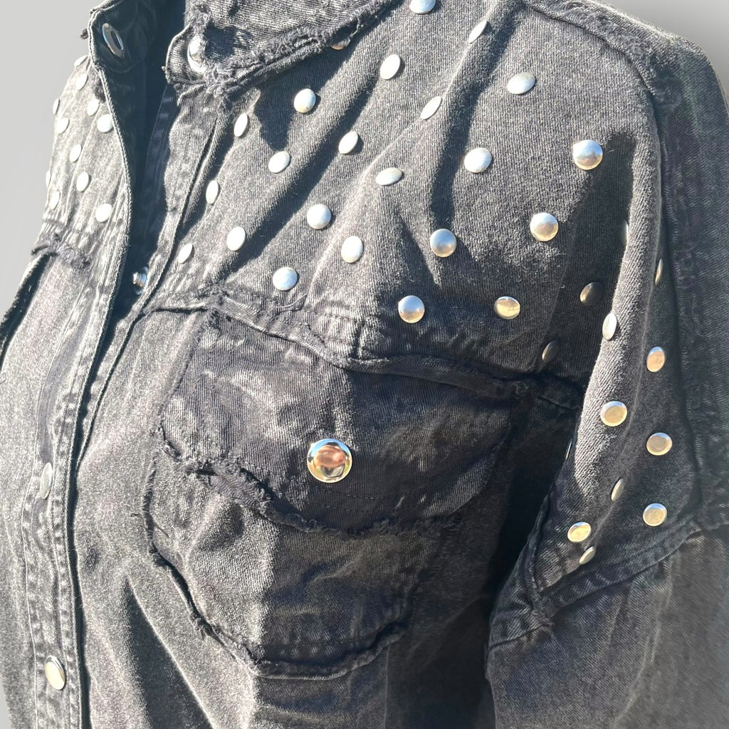Close-up of a black washed denim jacket decorated with silver metal studs and rivets, featuring a distressed pocket and raw hem detail.