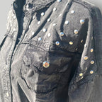 Close-up of a black washed denim jacket decorated with silver metal studs and rivets, featuring a distressed pocket and raw hem detail.