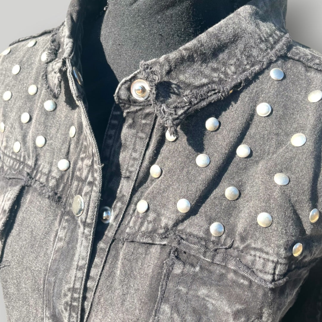 A close-up of a distressed black denim jacket with silver metal studs and a frayed collar, displayed on a black mannequin torso.