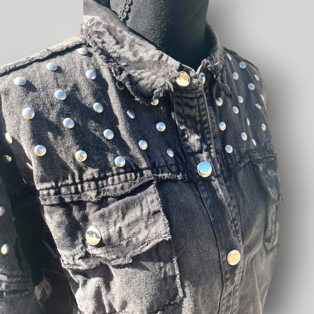 A close-up of a black washed denim jacket displayed on a black mannequin torso. The jacket features prominent silver-tone metal studs embellished across the chest and shoulders, with a frayed collar and button-front closure.