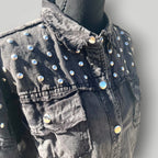 A close-up of a black washed denim jacket displayed on a black mannequin torso. The jacket features prominent silver-tone metal studs embellished across the chest and shoulders, with a frayed collar and button-front closure.