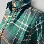 A close-up of a green plaid flannel shirt on a mannequin, featuring white and yellow intersecting stripes, gold buttons, and a dark collar.
